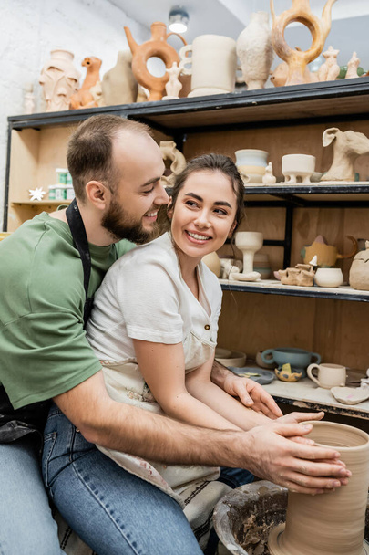 Positieve ambachtsman in schort maken klei vaas en kijken naar vriendje in de buurt aardewerk wiel in de studio - Foto, afbeelding