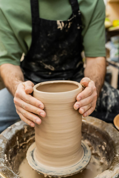Vista cortada do artesão borrado em avental moldando vaso de argila na roda de cerâmica no estúdio de cerâmica - Foto, Imagem