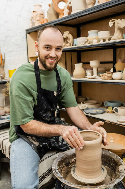 Glimlachend baard artisanaal in schort kijken naar camera en vormgeven klei vaas op aardewerk wiel in de studio - Foto, afbeelding