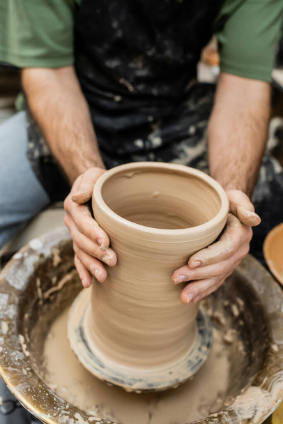 Vista recortada de alfarero borroso en delantal moldeado jarrón de arcilla en la rueda de cerámica en taller de cerámica - Foto, Imagen