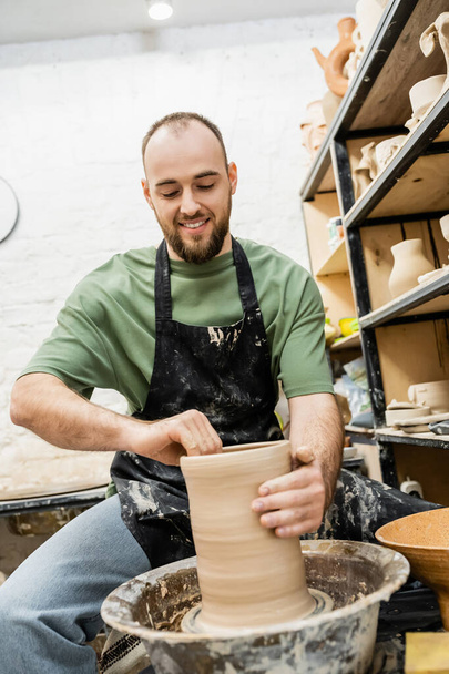 Usmívající se vousatý muž řemeslník v zástěře tvarování hliněné plastiky na keramické kolo v keramické dílně - Fotografie, Obrázek