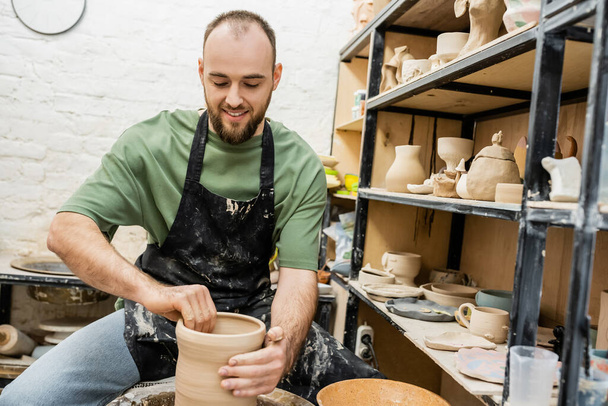Alegre barbudo artesano en delantal formando jarrón de arcilla en rueda de cerámica cerca de rack en taller - Foto, imagen