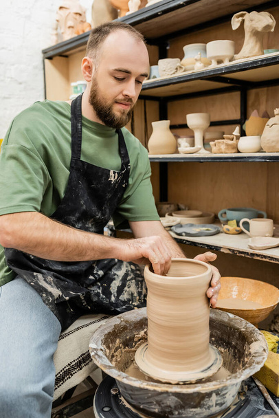 Artesano barbudo en delantal haciendo jarrón de barro sobre rueda de cerámica cerca de rack con esculturas en taller - Foto, imagen