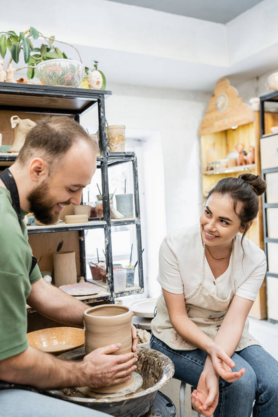 Joyful artisan in apron talking to boyfriend making clay vase on pottery wheel in ceramic workshop - Fotó, kép