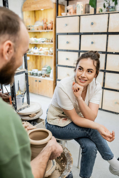 Positive craftsman in apron talking to girlfriend making clay vase on pottery wheel in workshop - Fotografie, Obrázek