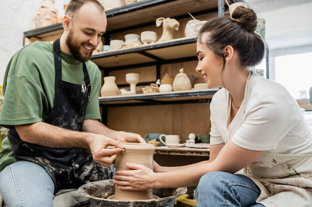 Artigiani romantici sorridenti in grembiuli che modellano insieme il vaso di argilla sulla ruota in ceramica nell'officina ceramica - Foto, immagini