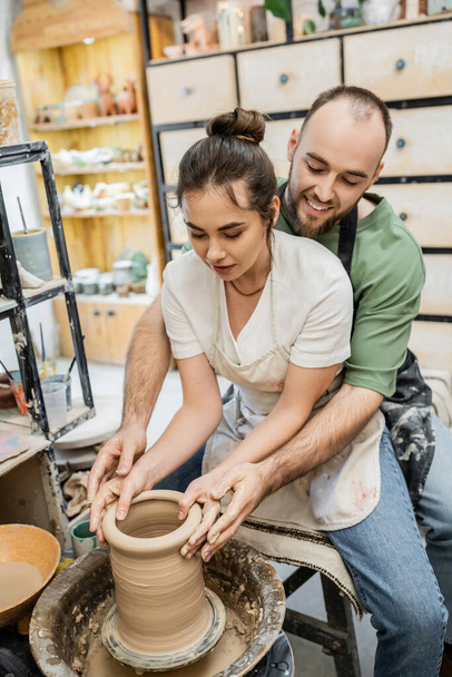 Smiling bearded artisan shaping clay vase together with girlfriend on pottery wheel in workshop - Foto, imagen