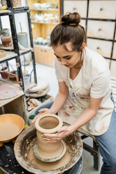 Morena artesana en delantal borroso haciendo jarrón de arcilla en la rueda de cerámica cerca del agua en el taller - Foto, Imagen