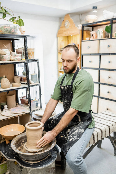 Бородатый ремесленник в грязном фартуке, формирующий глину на гончарном круге керамической студии - Фото, изображение