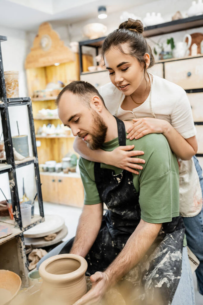 Alfarera sonriente en delantal abrazando novio trabajando con arcilla y rueda de cerámica en taller - Foto, Imagen