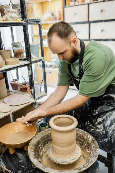 Bearded sculptor in apron washing hands in bowl with water near clay and pottery wheel in workshop - Photo, Image