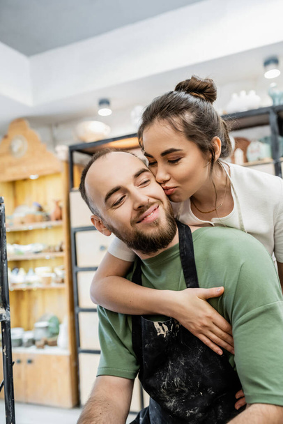 Mujer besar y abrazar novio artesanal en delantal mientras trabajan juntos en taller de cerámica - Foto, imagen