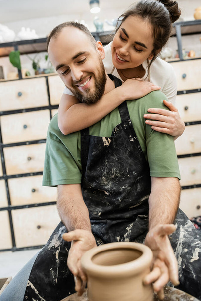 Smiling craftswoman embracing boyfriend in apron shaping clay vase on pottery wheel in studio - 写真・画像