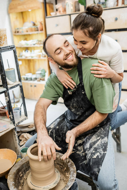 Handwerkerin in Schürze umarmt lächelnden Freund und formt Ton auf Töpferscheibe in Werkstatt - Foto, Bild
