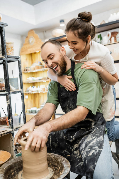 Веселий майстер в фартусі, який обіймає хлопця, роблячи глиняну вазу на керамічному колесі в майстерні - Фото, зображення
