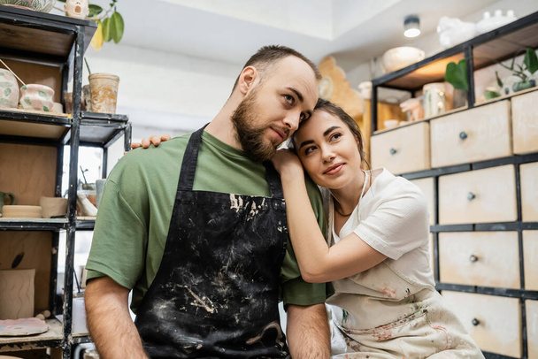 Craftswoman in apron hugging bearded boyfriend and looking away in ceramic studio at background - Фото, изображение