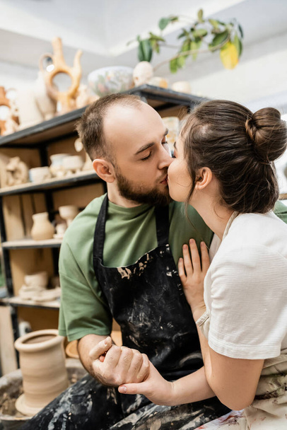 Craftsman in apron kissing and holding hand of girlfriend near blurred pottery wheel in workshop - Foto, Bild