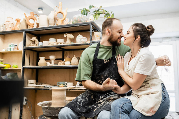 Vista lateral do casal de artesãos em aventais de mãos dadas e sorrindo perto da roda de cerâmica no estúdio - Foto, Imagem