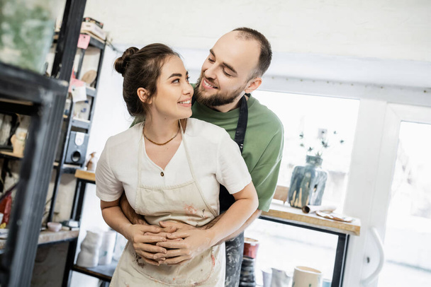 Smiling bearded artisan in apron hugging and looking at girlfriend in ceramic workshop - Foto, imagen