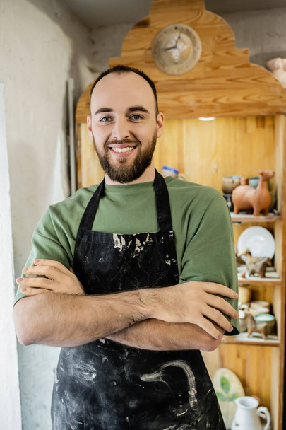 Cheerful bearded craftsman in apron crossing arms and looking at camera while standing in workshop - Photo, Image