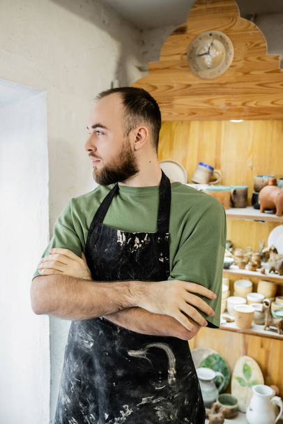 Brodatego rzemieślnika w fartuchu krzyżującym ramiona i odwracającego wzrok stojąc w warsztacie ceramicznym - Zdjęcie, obraz