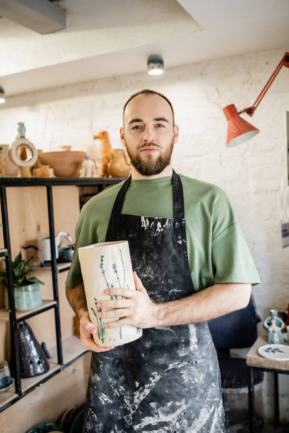 Artesano barbudo en delantal sosteniendo jarrón de cerámica mientras está de pie y mirando a la cámara en el taller - Foto, Imagen