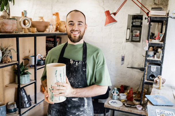 Glimlachend baard ambachtsman in schort met klei vaas en kijkend naar de camera in keramische werkplaats - Foto, afbeelding