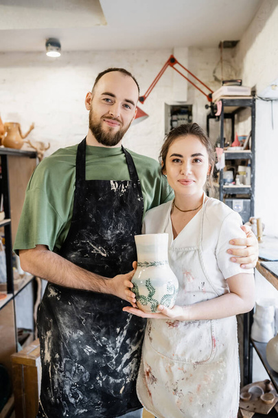 Lächelndes Handwerker-Paar in Schürzen umarmt und hält Tonvase in Keramik-Werkstatt im Hintergrund - Foto, Bild