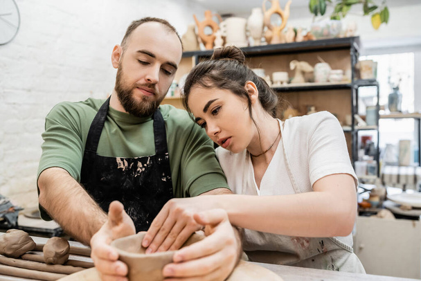 Artesana en delantal formando tazón de barro con novio y hablando mientras trabaja en taller de cerámica - Foto, Imagen