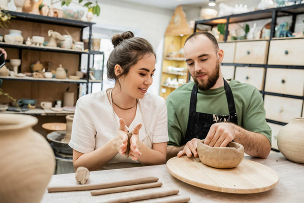 Smiling craftswoman in apron molding clay while boyfriend making bowl in ceramic workshop - Foto, imagen