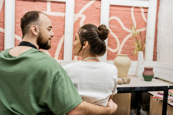 Бородатый мастер в фартуке обнимает подругу, сидя рядом с глиной в керамической мастерской - Фото, изображение