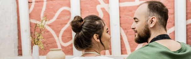 Brodaty rzemieślnik patrząc na brunetkę dziewczyna w pobliżu gliny w warsztacie garncarskim, sztandar - Zdjęcie, obraz