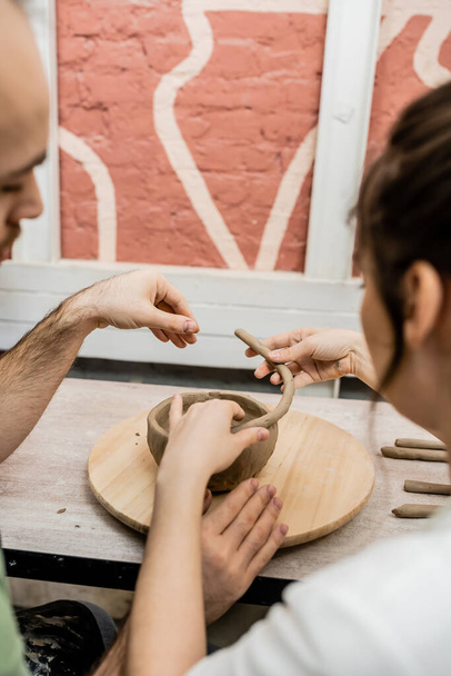 Pareja borrosa de artesanos moldeando tazón de arcilla en tablero de madera en el estudio de cerámica en el fondo - Foto, Imagen