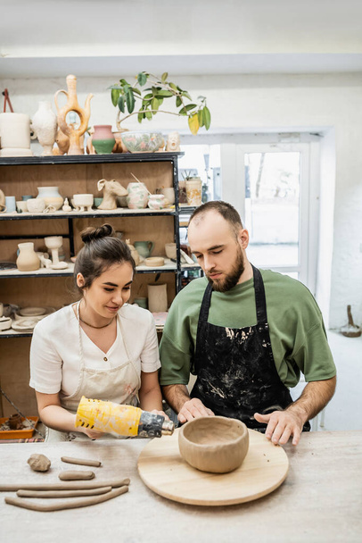 Smiling female artisan in apron drying ceramic bowl with heat gun near boyfriend in ceramic workshop - Foto, Imagem