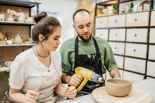 Craftsman in apron drying ceramic bowl with heat gun on wooden board near girlfriend in studio - Foto, immagini