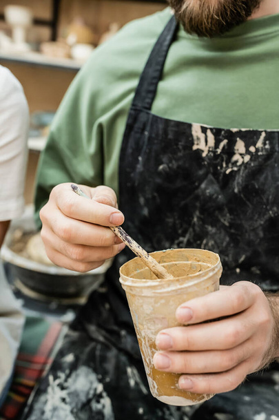 Cropped view of craftsman in apron holding clastic cup with dye and paintbrush in ceramic workshop - Photo, Image