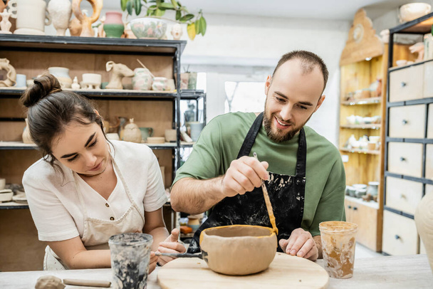 Sorridente artigiano colorare ciotola argilla vicino fidanzata in laboratorio di ceramica sullo sfondo - Foto, immagini