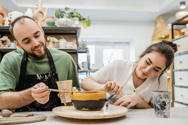 Artesanos alegres en delantales para colorear tazón de arcilla mientras trabajan juntos en taller de cerámica - Foto, imagen