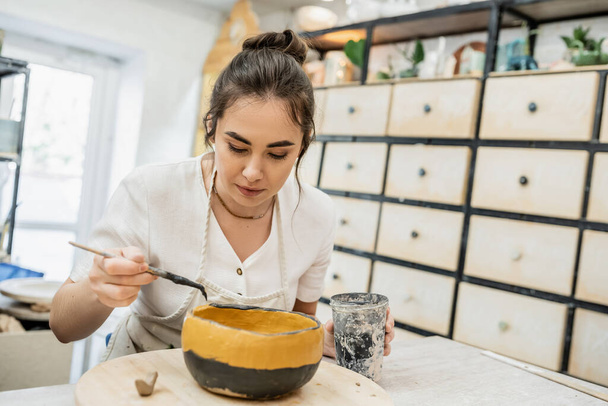 Morena artesana en delantal para colorear tazón de cerámica mientras trabaja en el estudio de cerámica en el fondo - Foto, Imagen