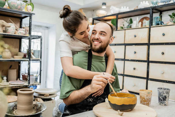 Joyful potter embracing boyfriend coloring clay bowl and working in ceramic studio at background - Foto, Imagem
