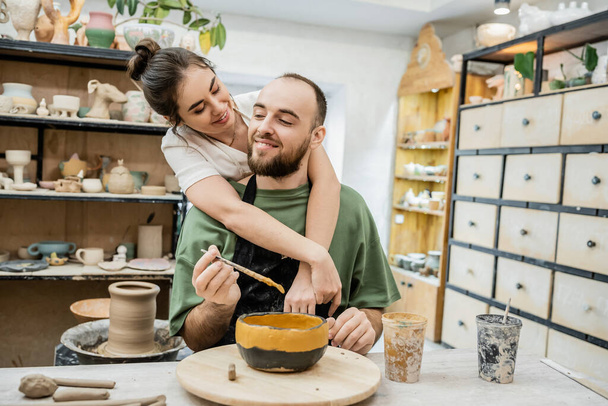 Smiling female potter hugging boyfriend coloring clay bowl while working in ceramic workshop - Photo, Image