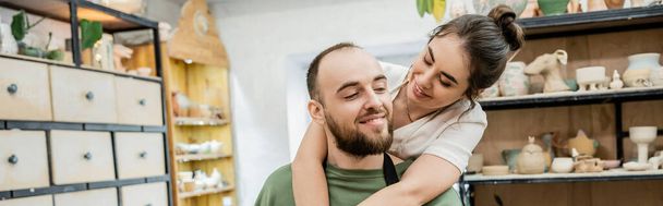 Artisanate souriante dans un tablier embrassant un petit ami barbu et travaillant dans un atelier de céramique, bannière - Photo, image