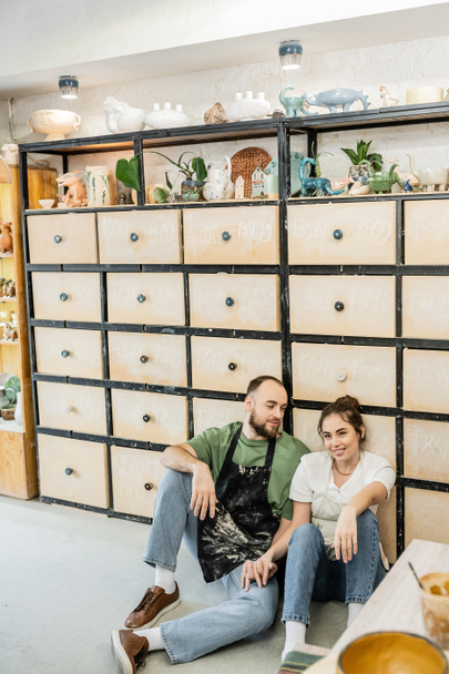 Craftsman in apron looking at smiling girlfriend sitting near cupboard in ceramic studio - Foto, Bild