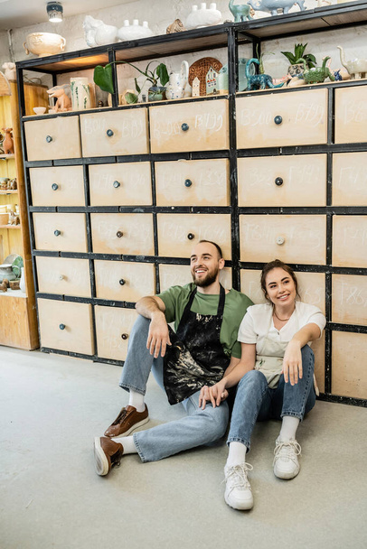 Zufriedenes Handwerker-Paar in Schürzen sitzt neben Schrank in Keramik-Werkstatt im Hintergrund - Foto, Bild