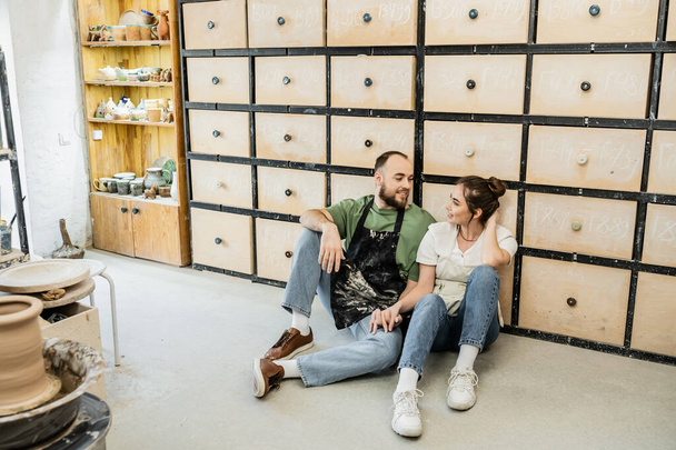 Couple joyeux de potiers dans des tabliers tenant la main tout en étant assis près du placard dans le studio de céramique - Photo, image