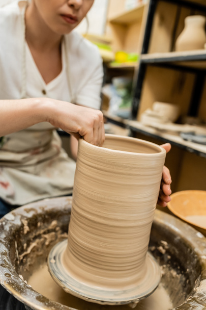 Vista recortada de la artesana en delantal moldeando arcilla sobre rueda de cerámica en taller borroso - Foto, imagen