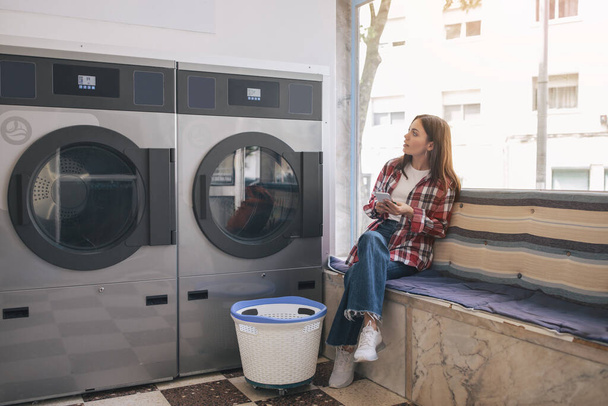 Young Woman In Casual Doing Laundry And Using Cell Phone, Sitting Waiting In Laundrette Room Near Washing Machines. Lady Booking Laundromat Via Mobile Application Posing Indoors - Foto, Imagem