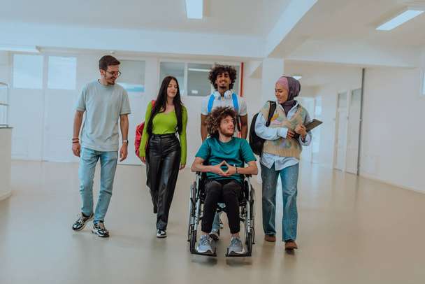 Nykyaikaisessa yliopistossa, monipuolinen ryhmä opiskelijoita, mukaan lukien afroamerikkalainen opiskelija ja hijabia käyttävä nainen, kävelevät yhdessä käytävällä, mukanaan pyörätuolilla sidottu kollegansa. - Valokuva, kuva