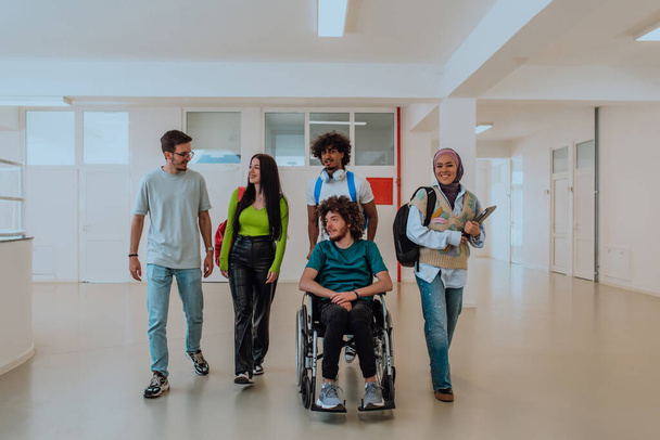 En una universidad moderna, un grupo diverso de estudiantes, incluyendo un estudiante afroamericano y una mujer que usa hiyab, caminan juntos en el pasillo, acompañados por su colega en silla de ruedas - Foto, imagen