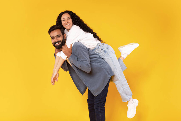 Happy Together. Romantic Middle Eastern Man Piggybacking His Cheerful Wife, Having Fun And Fooling Over Yellow Background In Studio, Couple Smiling At Camera Posing Joyfully - Photo, Image